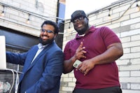 two men standing next to a laptop in front of a brick wall